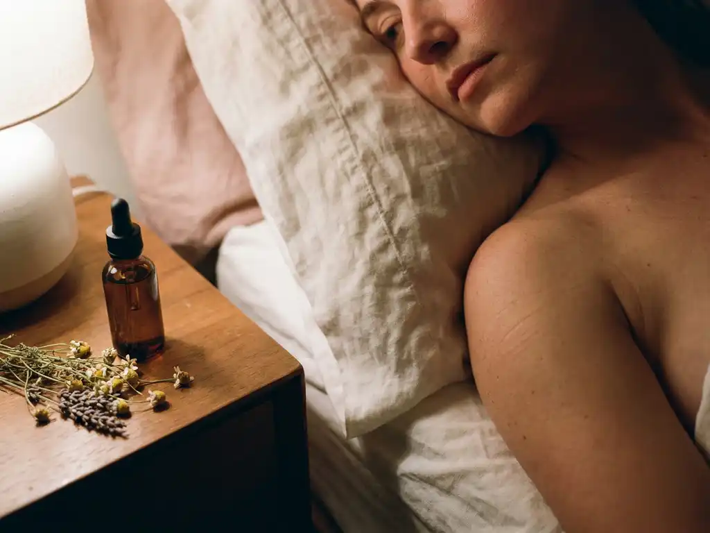 Vrouw rust op wit linnen kussen met zichtbare huidtextuur, naast een glazen flesje amber supplement en verse kruiden op het nachtkastje.